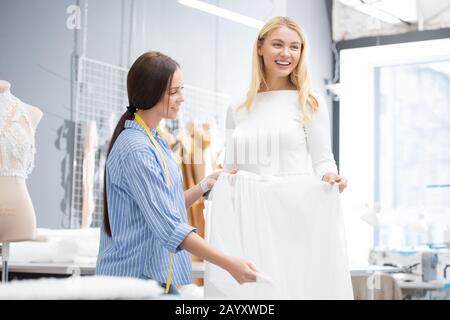 Happy Caucasian woman trying on wedding dress in modern bridal workshop, young seamstress helping her Stock Photo