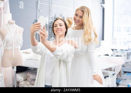 Horizontal portrait of two Caucasian women taking selfie shot on smartphone camera in wedding dress-making studio Stock Photo
