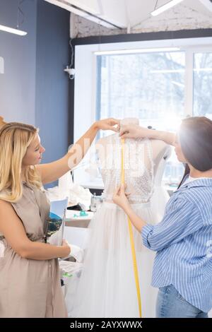 Two young bridal fashion designers working together on wedding gown in their workshop, vertical shot Stock Photo