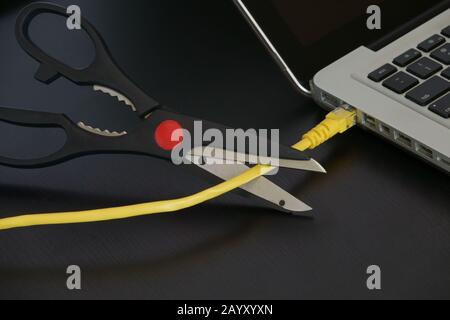 A pair of sharp scissors are shown just before cutting a yellow ethernet internet cable connected to a laptop computer. Stock Photo