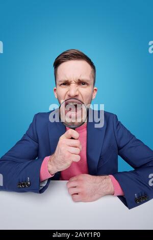Portrait of distraught young man in jacket sitting at table and screaming through magnifying glass against blue background Stock Photo