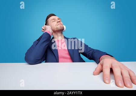 Relaxed young bearded man sitting with closed eyes at table and listening to inspiring music in headphones Stock Photo