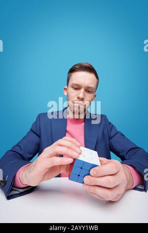 Serious young man in jacket sitting at table and solving cube puzzle while developing logic Stock Photo
