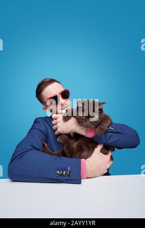 Young man in heart-shaped sunglasses sitting at table and holding displeased cat against blue background Stock Photo