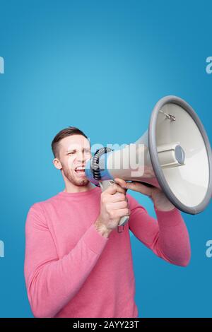 Young man with expressive face speaking announcement into megaphone against blue background Stock Photo