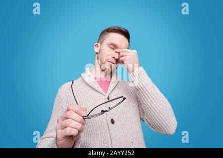 Tired young bearded man in cardigan rubbing bridge of nose and holding glasses against blue background Stock Photo