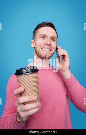 Smiling handsome young man with stubble drinking coffee from takeout cup and chatting by cellphone Stock Photo