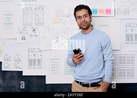 Horizontal medium portrait shot of young Caucasian man working in IT industry standing with smartphone looking at camera Stock Photo