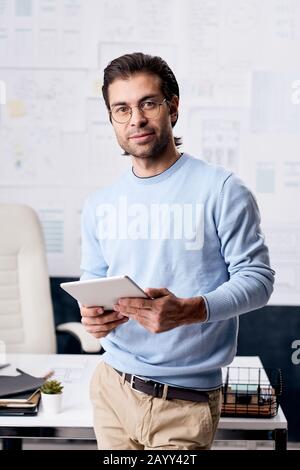 Vertical medium long portrait of modern handsome businessman wearing eyeglasses holding tablet PC looking at camera Stock Photo