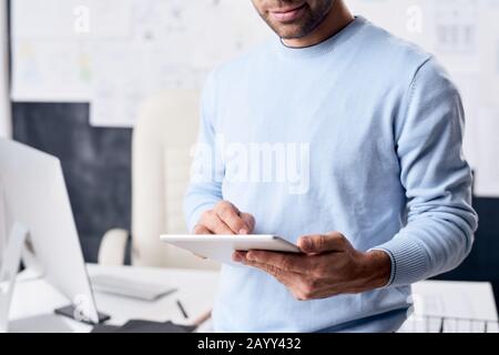 Horizontal shot of unrecognizable young man wearing light blue sweater standing in office using tablet PC Stock Photo