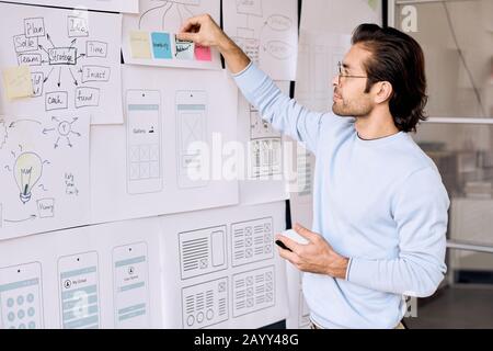 Horizontal shot of IT specialist working on project using sheets of paper with schemes and notes fixed on blackboard Stock Photo
