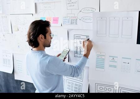 Horizontal shot of handsome young man wearing eyeglasses holding smartphone making notes on paper with software sketch Stock Photo