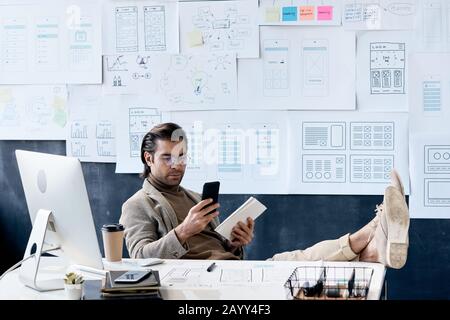 Stylish Caucasian man sitting relaxed at his office desk with feet on it holding smartphone at tablet PC Stock Photo