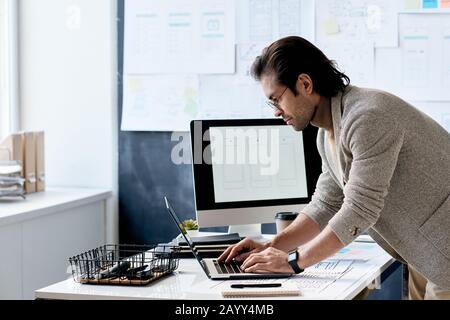 Horizontal side view shot of handsome man wearing eyeglasses standing at office desk working on laptop Stock Photo