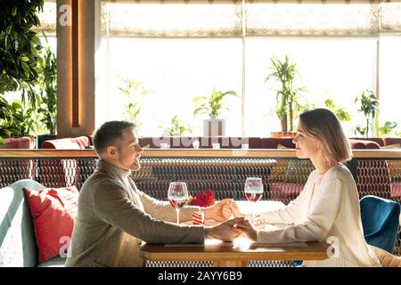 Horizontal side view shot of happy man and woman in love sitting in front of each other in restaurant holding their hands Stock Photo