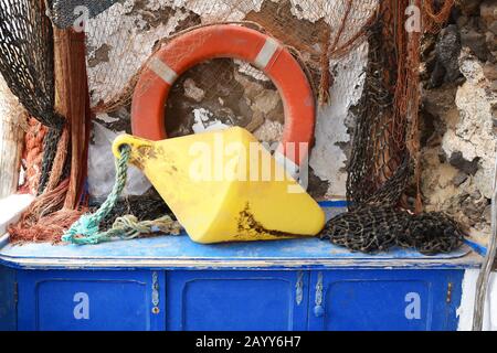 Collection of brightly coloured fishing equipment on blue wooden chest with stone wall. Gear includes weight, life saver ring, nets and rope. Stock Photo