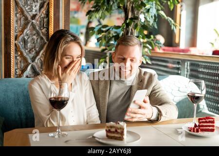 Horizontal shot of young man and woman sitting together at cafe table watching something funny on smartphone and laughing Stock Photo