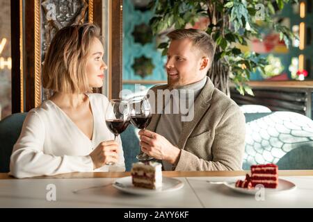 Handsome Caucasian man and beautiful woman sitting together at cafe table clinking glasses with red wine looking at each other Stock Photo
