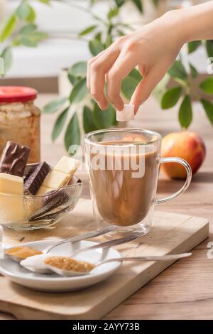 The boy's hand is holding a sugar cube over a glass of tea or coffee with milk. Different types of sweets and sugar lie on the table. Stock Photo