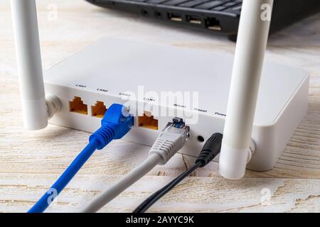 Close-up of network cables connected to a white Wi-Fi wireless router near laptop on a white wooden table. Wlan router with internet cables plugged in Stock Photo