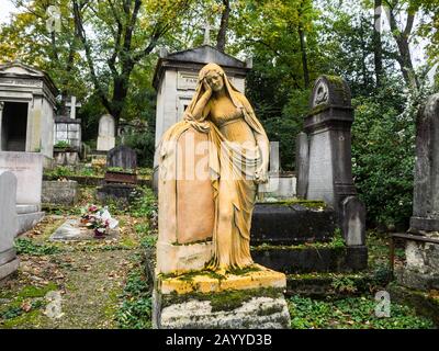 Old tombstone covered with musk with a statue of a young woman crying over a plaque in a path under the trees of monumental cementery Stock Photo