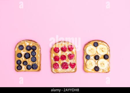 Top view of peanut butter sandwiches or toasts with banana, raspberry and blueberry on soft pink background with copy space. Healthy eating concept Stock Photo