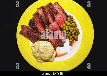Medium-rare homemade steak with peas and cream sauce on yellow plate isolated on black Stock Photo