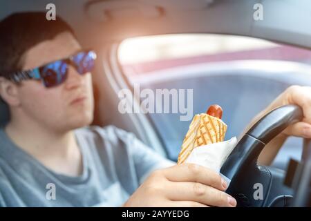 Adult male caucasian person holding hot dog fast food snack while driving car on sunny day. Men eating food while driving steering wheel. Distracted Stock Photo