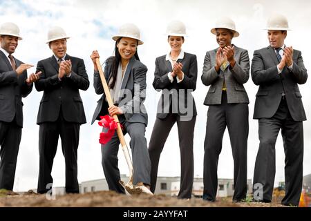 Smiling middle-aged businesswoman performs the groundbreaking ceremony of a new development as her colleagues look on happily and applaud. Stock Photo