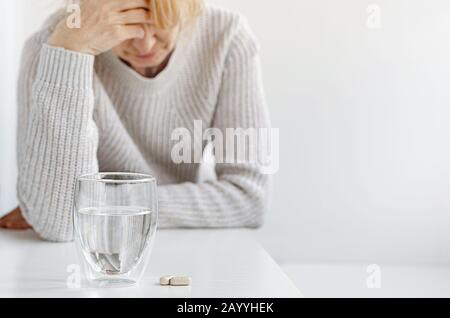 Elderly woman holding hand on her head. Selective focus on pills and glass of water. Headache and stress concept Stock Photo