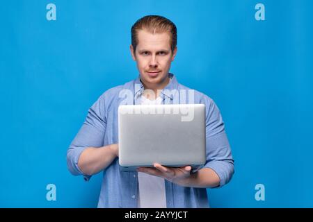 A man in a blue shirt holds a laptop and looks at the camera. Programmer concept working on troubleshooting. Close up. Stock Photo
