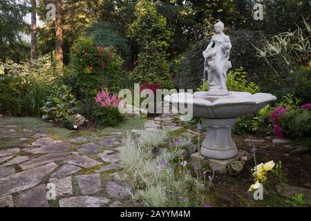 Flagstone path, water fountain and borders with ornamental grass plants, yellow Rosa ‘Flower Carpet' - Roses, Filipendula purpurea ‘Elegans'. Stock Photo