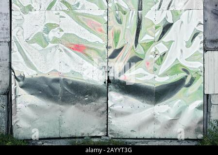 Silver metal sheets are nailed onto a garage door, simply closed with a small chain. The metal sheets are wavy and reflecting green and red colors. Stock Photo