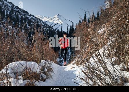 Old man with grey beard and red jacket is running near on the trail at mountains in winter time. Skyrunning and trailrunning outdoor activity concept.