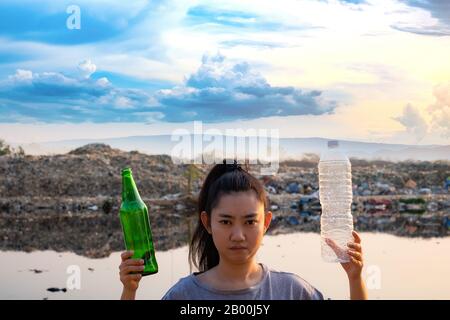 A woman stands hand holding green glass bottle and clear plastic bottle at mountain large garbage background, These waste come from urban and industri Stock Photo