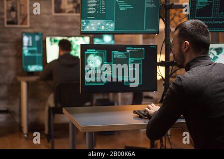 Back view of hackers working on making a dangerous malware on computers with multiple screens. Stock Photo