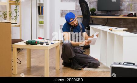 Male worker talking on the phone while assembles white furniture in new home. Electric drill. Stock Photo