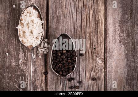 Vintage effect image of tarnished silver spoons filled with salt crystals and black peppercorns, over rough wood background. Stock Photo