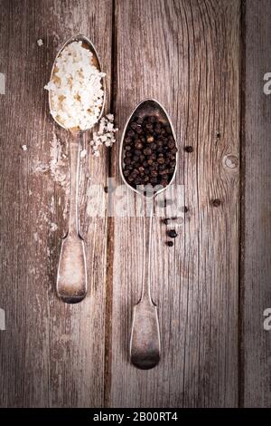 Vintage effect image of tarnished silver spoons filled with salt crystals and black peppercorns, over rough wood background. Stock Photo