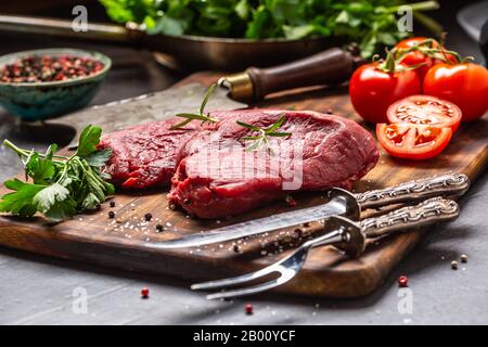 Two beef rump steaks with herbs tomatoes salt and spices on butcher board. Stock Photo