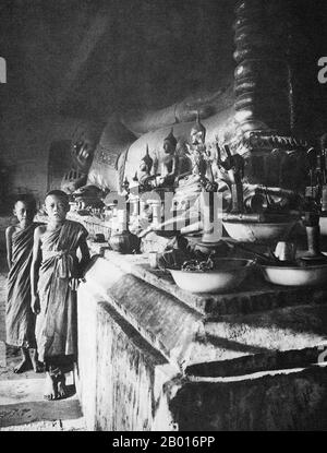 Thailand: Two novice monks stand next to a reclining Buddha, Wat Phra That Haripunchai, Lamphun, c. 1920.  Wat Phra That Haripunchai was founded in 1044 by King Athitayarat of Haripunchai on the site of Queen Chamathewi's (Chama Thewi or Chamadevi) royal palace. Legend has it that the queen's personal quarters are enclosed in the main 46-metre high Lan Na-style chedi, covered in copper plates and topped by a gold umbrella or plee.   Lamphun was the capital of the small but culturally rich Mon Kingdom of Haripunchai from about 750 CE to the time of its conquest by King Mangrai in 1281. Stock Photo