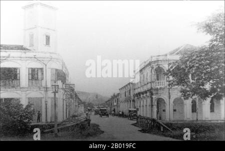 Thailand: Street scene with vintage car and Sino-Portuguese shophouses in Phuket, early 20th century.  Phuket, formerly known as Talang and, in Western sources, Junk Ceylon (a corruption of the Malay Tanjung Salang, i.e. 'Cape Salang'), is one of the southern provinces (changwat) of Thailand. Neighbouring provinces are (from north clockwise) Phang Nga and Krabi, but as Phuket is an island there are no land boundaries.  Phuket, which is approximately the size of Singapore, is Thailand’s largest island. The island is connected to mainland Thailand by two bridges. Stock Photo