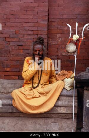 Nepal: A sadhu with his trident and smoking hashish, Kathmandu.  They are known, variously, as sadhus (saints, or 'good ones'), yogis (ascetic practitioners), fakirs (ascetic seeker after the Truth) and sannyasins (wandering mendicants and ascetics). They are the ascetic – and often eccentric – practitioners of an austere form of Hinduism. Sworn to cast off earthly desires, some choose to live as anchorites in the wilderness. Others are of a less retiring disposition, especially in the towns and temples of Nepal's Kathmandu Valley. Stock Photo