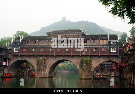 China: Mist hangs over Hong Qiao Bridge, Fenghuang's famed covered bridge.  Fenghuang is Chinese for Phoenix and refers to the mythical sacred firebird that can be found in the mythologies of the Persians, Greeks, Romans, Egyptians, Chinese, and (according to Sanchuniathon) the Phoenicians.  Legend suggests that two phoenixes on discovering the town hovered overhead for some considerable time before reluctantly flying away.  Fenghuang town is a well-preserved ancient town supposedly dating back to 248 BC. It is home to the Miao and Tujia minorities. Stock Photo