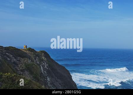 Small sigluestea of couple and dog standing on the cliff wathcing to waves on the ocean Stock Photo
