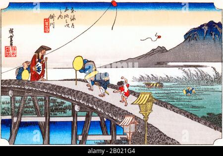 Kakegawa: Travellers crossing a high trestle-bridge over the Kake River, two of them peering into the water below, and behind a small boy watching a kite up in the air, while beyond another, with broken string, flutters to earth. Peasants transplanting rice in the flooded fields, and in the distance Mount Akiba rising above the mists. A shrine standing on the top of a mountain near this station attracted worshippers from all over Japan who came to pray for protection against the calamity of fire. An old couple of travellers are struggling against a strong wind as they toil to cross the arched Stock Photo