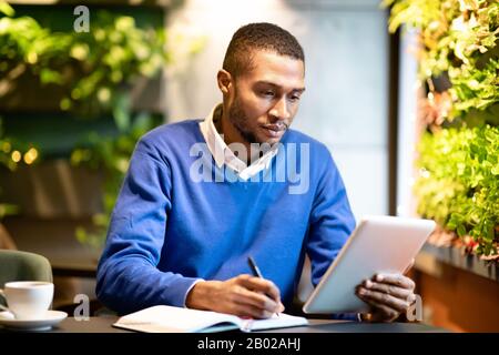 Black business guy taking notes in notebook holding tablet Stock Photo