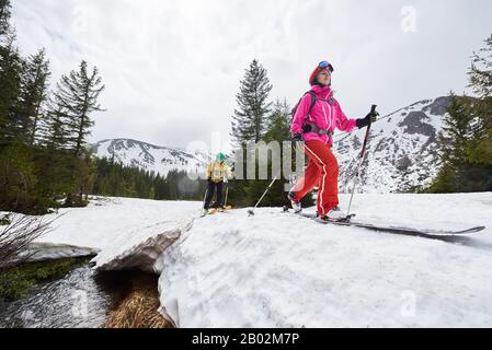 Skiing off-piste on foothill plain. Low angle view of skiers young woman and man during ski touring over flowing river under snowdrift. Activities in mountains under grey sky. Trees on background Stock Photo