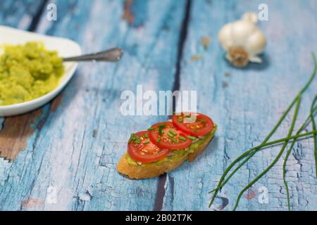 An open-faced sandwich made with baguette, avocado spread, slices of cherry tomato, and diced chives, chives green stalks are visible next to it. Stock Photo