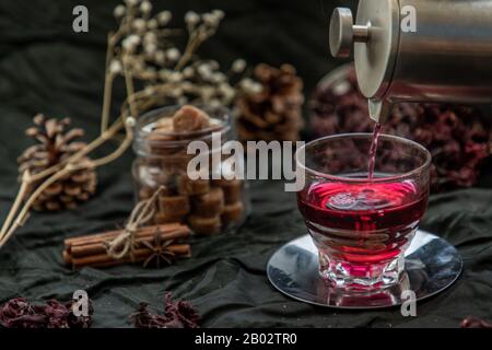 Roselle tea (Jamaica sorrel, Rozelle or hibiscus sabdariffa ) is poured from a kettle into glass cup  with dry roselle and brown cane sugar cube. Heal Stock Photo